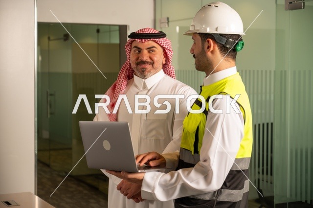 عقد صفقات عمل جديدة، التفاهم والتعاون بين الزملاء، ايماءات السعادة والسرور، تبادل الخبرات والمعلومات لتحقيق الاهداف، مهندس عربي خليجي سعودي يرتدي سترة العمل وخوذة الحماية البيضاء يقف مع زميله، مفهوم الهندسة والإنشاءات المعمارية، مشاريع إعمارية سعودية وطنية