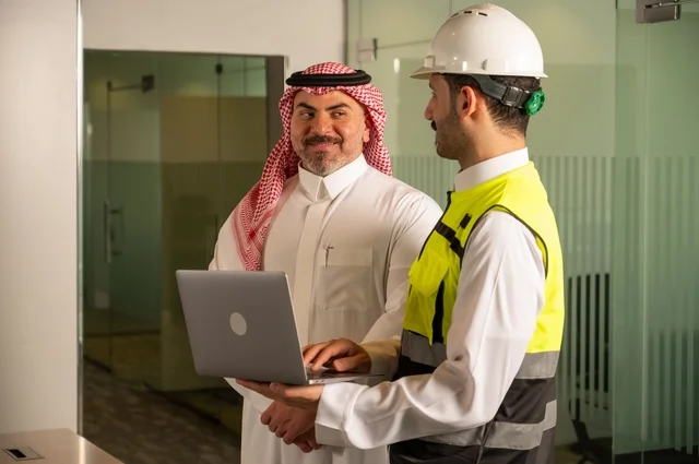 عقد صفقات عمل جديدة، التفاهم والتعاون بين الزملاء، ايماءات السعادة والسرور، تبادل الخبرات والمعلومات لتحقيق الاهداف، مهندس عربي خليجي سعودي يرتدي سترة العمل وخوذة الحماية البيضاء يقف مع زميله، مفهوم الهندسة والإنشاءات المعمارية، مشاريع إعمارية سعودية وطنية