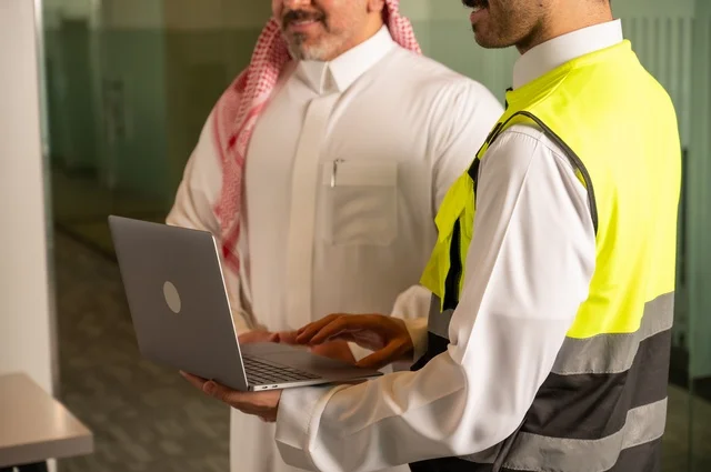 Understanding and cooperation among colleagues, making new business deals, exchanging experiences and information to achieve goals, a close-up image of a Saudi Gulf Arab engineer wearing a work jacket and a white safety helmet standing with a colleague, the concept of engineering and architectural construction, national Saudi construction projects.