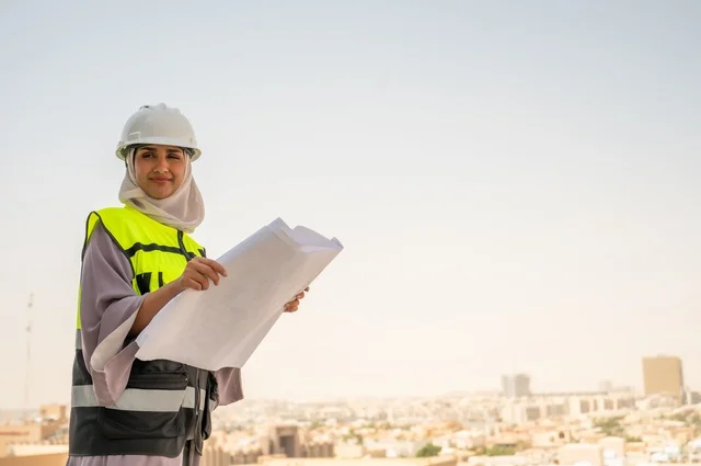 Construction project, side view of a Saudi Arabian Gulf female architect holding a building plan in her hand, a smiling hijab-wearing construction engineer dressed in work safety gear, a work helmet, formal work attire, female engineering professions and jobs, background of towers and skyscrapers.