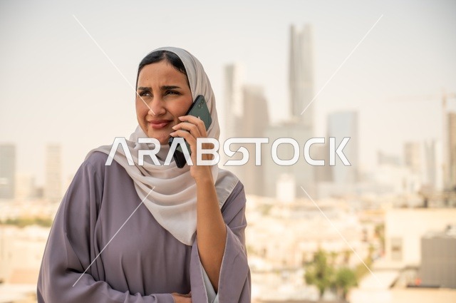 استخدام جهاز تقني حديث ومتطور، صورة مقربة لامرأة عربية خليجية عربية سعودية ترتدي العباءة والحجاب تقوم باستخدام الهاتف المحمول،التواصل مع العملاء عن بعد، دمج التكنولوجيا بالعمل، إنجاز المهام المطلوبة اليومية، مهنة ووظيفة نسائية مكتبية