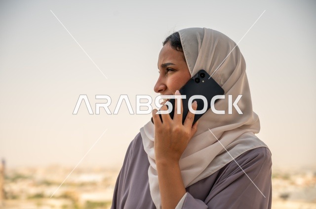 إجراء اتصال هاتفي عبر الجوال, استخدام جهاز تقني حديث ومتطور في العمل, مهنة ووظيفة مكتبية في السعودية, امرأة عربية خليجية سعودية محجبة ترتدي العباءة تقف في الهواء الطلق وتمسك الهاتف المحمول بيدها,التواصل مع العملاء عن بعد