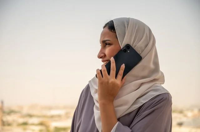 Making a phone call via mobile, using a modern and advanced technical device at work, a profession and office job in Saudi Arabia, an Arab Gulf Saudi woman wearing a hijab and an abaya standing outdoors holding a mobile phone in her hand, communicating with clients remotely.
