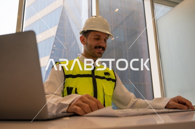 الإعمار بسواعد أبناء الوطن،  مفهوم الهندسة والإنشاءات، مهندس عربي خليجي سعودي يرتدي سترة وخوذة الحماية يجلس على المكتب يتأمل المخطط الهندسي ويدون الملاحظات، تدقيق ومراجعة الخطط الهندسية، دراسة أساسيات المشروع، إكمال رسم مخطوطات البناء