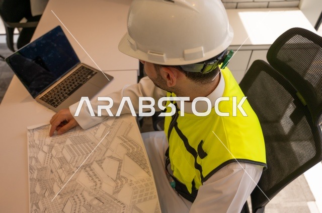 صورة علوية مقربة لمهندس عربي خليجي سعودي يرتدي سترة وخوذة الحماية يجلس على المكتب يتأمل المخطط الهندسي ويدون الملاحظات،الإعمار بسواعد أبناء الوطن،  مفهوم الهندسة والإنشاءات، تدقيق ومراجعة الخطط الهندسية، دراسة أساسيات المشروع، إكمال رسم مخطوطات البناء