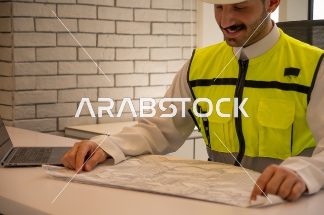 الإعمار بسواعد أبناء الوطن،  مفهوم الهندسة والإنشاءات، مهندس عربي خليجي سعودي يرتدي سترة وخوذة الحماية يجلس على المكتب يتأمل المخطط الهندسي ويون الملاحظات، تدقيق ومراجعة الخطط الهندسية، دراسة أساسيات المشروع، إكمال رسم مخطوطات البناء