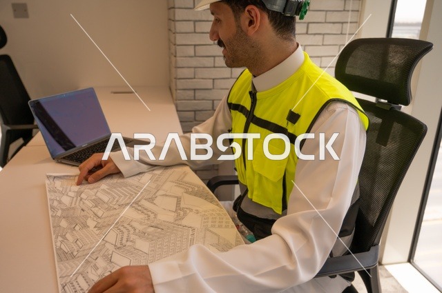 إكمال رسم مخطوطات البناء، مهندس عربي خليجي سعودي يرتدي سترة وخوذة الحماية يجلس على المكتب يتأمل المخطط الهندسي ويدون الملاحظات،الإعمار بسواعد أبناء الوطن،  مفهوم الهندسة والإنشاءات، تدقيق ومراجعة الخطط الهندسية، دراسة أساسيات المشروع