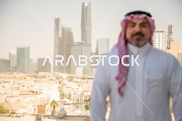 الاناقة والاهتمام بالمظهر الخارجي. صورة مقربة لرجل عربي خليجي سعودي مبتسم يرتدي الثوب والشماغ التقليدي ينظر الى الكاميرا بايماءات السرور والسعادة. الخروج في الهواء الطلق،  مفهوم الرجولة والثقة بالنفس، خلفية أبنية