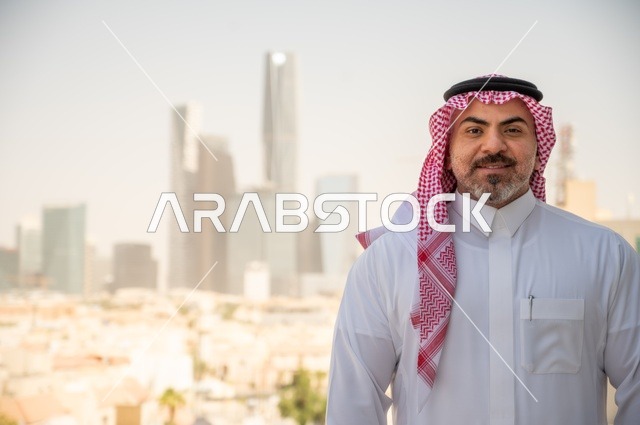 مفهوم الرجولة والثقة بالنفس، الاناقة والاهتمام بالمظهر الخارجي. صورة مقربة لرجل عربي خليجي سعودي مبتسم يرتدي الثوب والشماغ التقليدي ينظر الى الكاميرا بايماءات السرور والفخر. الخروج في الهواء الطلق، خلفية أبراج وناطحات سحاب في الرياض