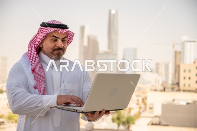 مفهوم تسهيل الاعمال والقدرة على إنجاز المهام، استخدام التقنيات الحديثة والأجهزة المتطورة ، رجل عربي خليجي سعودي يرتدي الثوب التقليدي يمسك بيده الحاسب المحمول لمتابعة المهام اليومية، خلفية الابراج وناطحات السحاب في الرياض