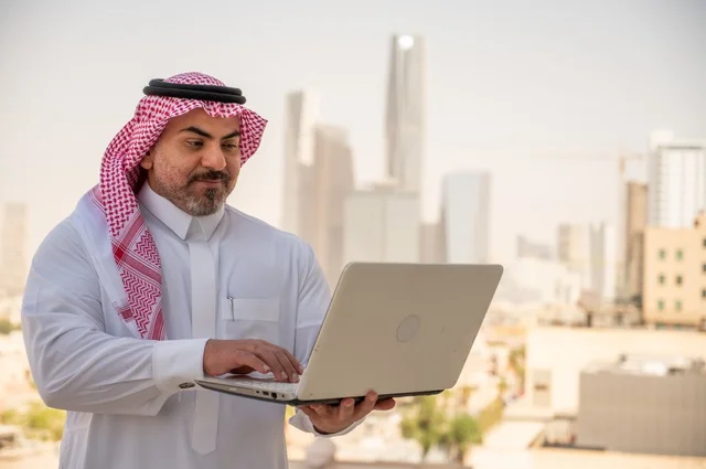 مفهوم تسهيل الاعمال والقدرة على إنجاز المهام، استخدام التقنيات الحديثة والأجهزة المتطورة ، رجل عربي خليجي سعودي يرتدي الثوب التقليدي يمسك بيده الحاسب المحمول لمتابعة المهام اليومية، خلفية الابراج وناطحات السحاب في الرياض