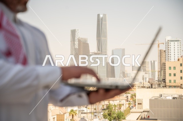 مهارة الأيدي السريعة في استخدام الأجهزة المتطورة في الحياة العملية، مفهوم تسهيل الاعمال والقدرة على إنجاز المهام، صورة جانبية مقربة لايدي رجل عربي خليجي سعودي يرتدي الثوب التقليدي يمسك بيده الحاسب المحمول لمتابعة المهام اليومية، خلفية الابراج وناطحات السحاب في الرياض