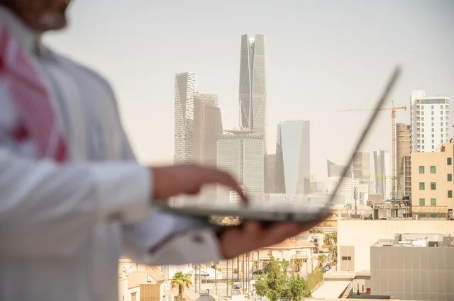 مهارة الأيدي السريعة في استخدام الأجهزة المتطورة في الحياة العملية، مفهوم تسهيل الاعمال والقدرة على إنجاز المهام، صورة جانبية مقربة لايدي رجل عربي خليجي سعودي يرتدي الثوب التقليدي يمسك بيده الحاسب المحمول لمتابعة المهام اليومية، خلفية الابراج وناطحات السحاب في الرياض