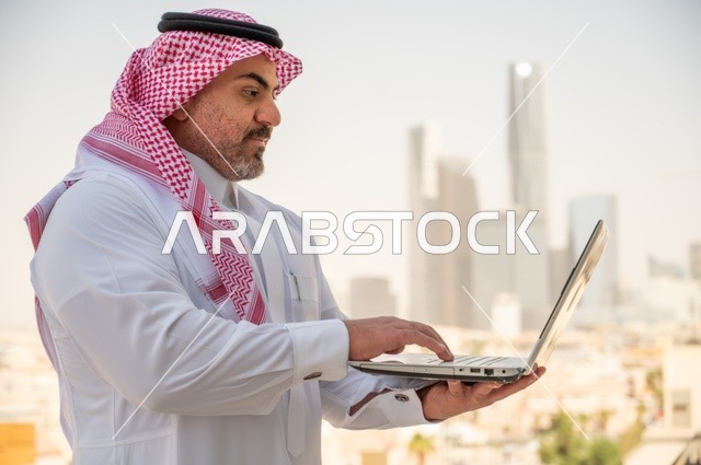 استخدام التقنيات الحديثة والأجهزة المتطورة، مفهوم تسهيل الاعمال والقدرة على إنجاز المهام، صورة جانبية مقربة لرجل عربي خليجي سعودي يرتدي الثوب التقليدي يمسك بيده الحاسب المحمول لمتابعة المهام اليومية، خلفية الابراج وناطحات السحاب في الرياض
