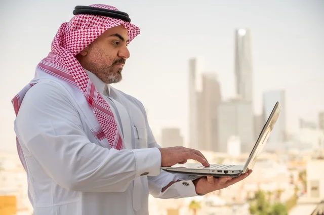 استخدام التقنيات الحديثة والأجهزة المتطورة، مفهوم تسهيل الاعمال والقدرة على إنجاز المهام، صورة جانبية مقربة لرجل عربي خليجي سعودي يرتدي الثوب التقليدي يمسك بيده الحاسب المحمول لمتابعة المهام اليومية، خلفية الابراج وناطحات السحاب في الرياض