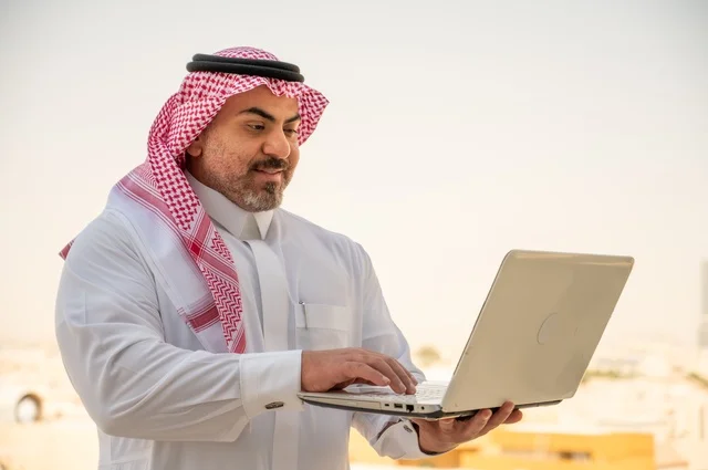 The use of modern technologies and advanced devices, the concept of facilitating work and the ability to accomplish tasks, a close-up side image of a Saudi Arabian Gulf man wearing traditional attire holding a laptop in his hand to follow up on daily tasks, with a background of towers and skyscrapers in Riyadh.