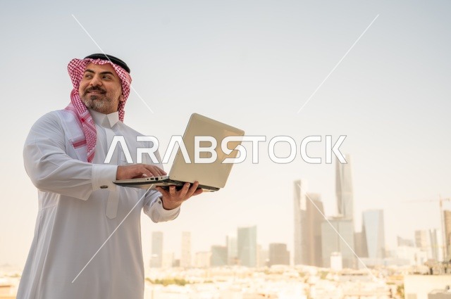 دمج التقنيات الحديثة والأجهزة المتطورة في الحياة العملية، مفهوم تسهيل الاعمال والقدرة على إنجاز المهام، صورة جانبية لرجل عربي خليجي سعودي يرتدي الثوب التقليدي يمسك بيده الحاسب المحمول لمتابعة المهام اليومية، خلفية الابراج وناطحات السحاب في الرياض