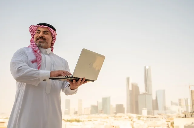 دمج التقنيات الحديثة والأجهزة المتطورة في الحياة العملية، مفهوم تسهيل الاعمال والقدرة على إنجاز المهام، صورة جانبية لرجل عربي خليجي سعودي يرتدي الثوب التقليدي يمسك بيده الحاسب المحمول لمتابعة المهام اليومية، خلفية الابراج وناطحات السحاب في الرياض