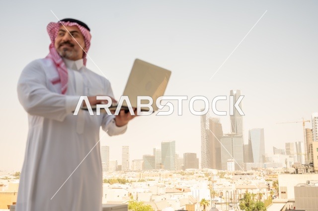 مفهوم تسهيل الاعمال والقدرة على إنجاز المهام، استخدام التقنيات الحديثة والأجهزة المتطورة ، رجل عربي خليجي سعودي يرتدي الثوب التقليدي يمسك بيده الحاسب المحمول لمتابعة المهام اليومية، خلفية الابراج وناطحات السحاب في الرياض