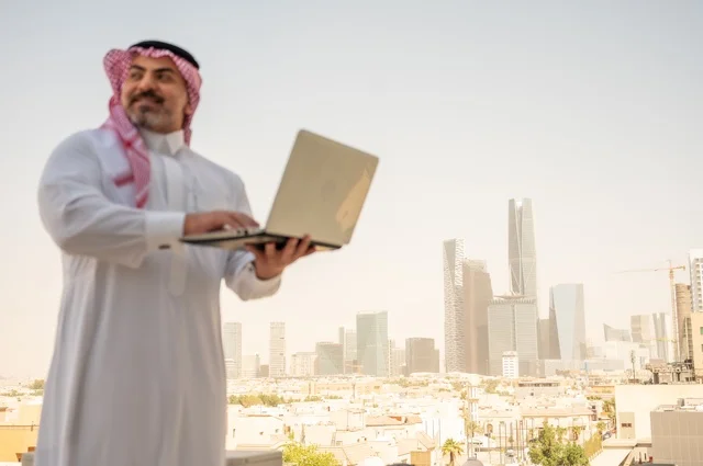 مفهوم تسهيل الاعمال والقدرة على إنجاز المهام، استخدام التقنيات الحديثة والأجهزة المتطورة ، رجل عربي خليجي سعودي يرتدي الثوب التقليدي يمسك بيده الحاسب المحمول لمتابعة المهام اليومية، خلفية الابراج وناطحات السحاب في الرياض