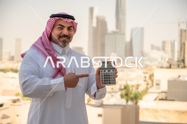 رجل عربي خليجي سعودي يرتدي الثوب التقليدي يمسك بيده آلة حاسبة لإجراء عمليات الحساب، مفهوم تسهيل الاعمال والقدرة على إنجاز المهام، استخدام التقنيات الحديثة والأجهزة المتطورة ،خلفية الابراج وناطحات السحاب في الرياض