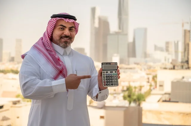 رجل عربي خليجي سعودي يرتدي الثوب التقليدي يمسك بيده آلة حاسبة لإجراء عمليات الحساب، مفهوم تسهيل الاعمال والقدرة على إنجاز المهام، استخدام التقنيات الحديثة والأجهزة المتطورة ،خلفية الابراج وناطحات السحاب في الرياض