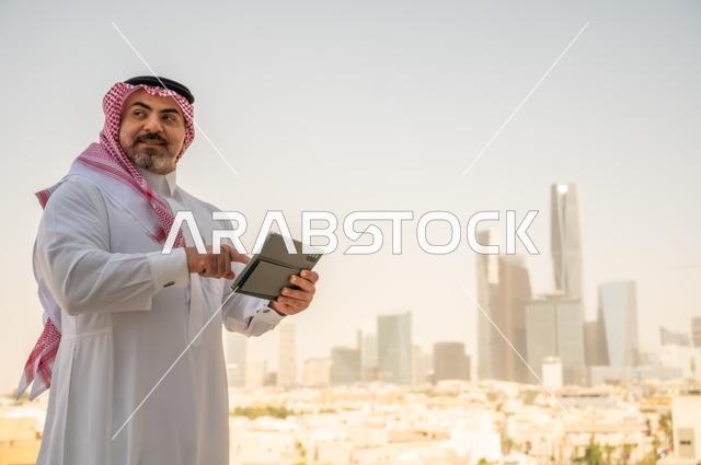 تصفح مواقع التواصل الاجتماعي، جهاز تقني حديث ومتطور، رجل عربي خليجي سعودي يرتدي الثوب والشماغ التقليدي يقوم باستخدام جهاز التابلت اللوحي، إنجاز العمل عن بعد، خلفية الأبراج وناطحات السحاب في مدينة الرياض