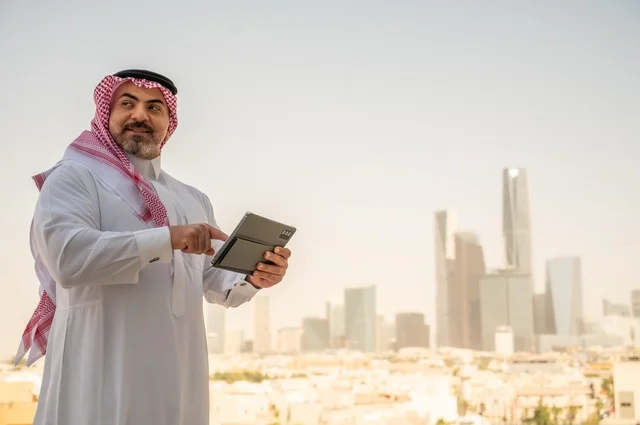 تصفح مواقع التواصل الاجتماعي، جهاز تقني حديث ومتطور، رجل عربي خليجي سعودي يرتدي الثوب والشماغ التقليدي يقوم باستخدام جهاز التابلت اللوحي، إنجاز العمل عن بعد، خلفية الأبراج وناطحات السحاب في مدينة الرياض