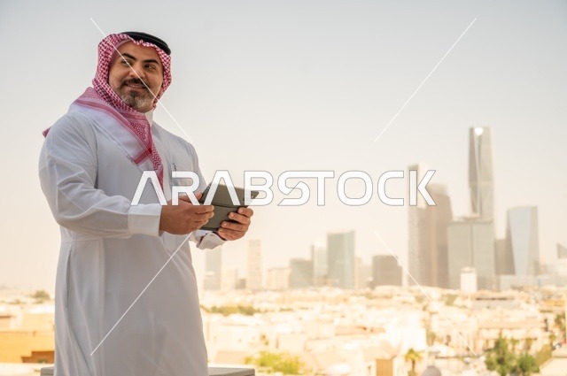 A modern and advanced technical device, browsing social media, an Arab Gulf Saudi man wearing traditional thobe and shemagh using a tablet device, accomplishing work remotely, with a background of towers and skyscrapers in the city of Riyadh.
