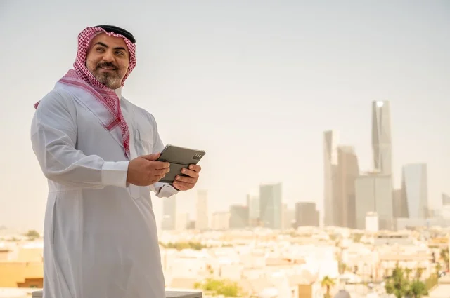 A modern and advanced technical device, browsing social media, an Arab Gulf Saudi man wearing traditional thobe and shemagh using a tablet device, accomplishing work remotely, with a background of towers and skyscrapers in the city of Riyadh.