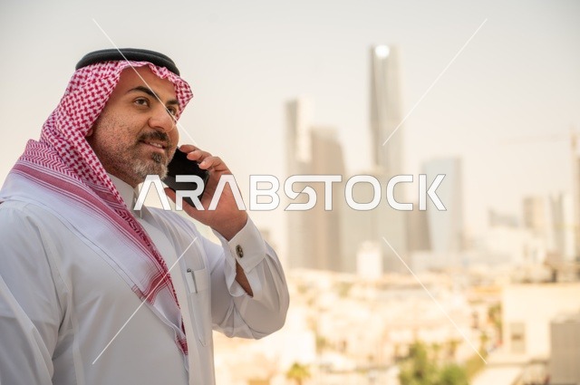 صورة مقربة لرجل عربي خليجي سعودي يرتدي الثوب والشماغ التقليدي يجري مكالمة هاتفية باستخدام المحمول، إنجاز العمل عن بعد،  نمو وتطور المجال الهندسي، جهاز تقني حديث ومتطور، تصفح مواقع التواصل الاجتماعي، خلفية الأبراج وناطحات السحاب في مدينة الرياض