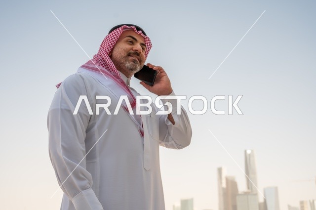 نمو وتطور المجال الهندسي، جهاز تقني حديث ومتطور، تصفح مواقع التواصل الاجتماعي، رجل عربي خليجي سعودي يرتدي الثوب والشماغ التقليدي يجري مكالمة هاتفية باستخدام المحمول، إنجاز العمل عن بعد، خلفية الأبراج وناطحات السحاب في مدينة الرياض