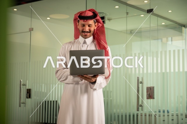 إدارة شؤون الأعمال وتنظيمها، استخدام جهاز تقني حديث ومتطور، مهنة ووظيفة ادارية مكتبية، رجل عربي خليجي سعودي يرتدي الثوب التقليدي يقوم بالعمل على الحاسوب المحمول داخل المكتب، الشركات والمكاتب في المملكة العربية السعودية