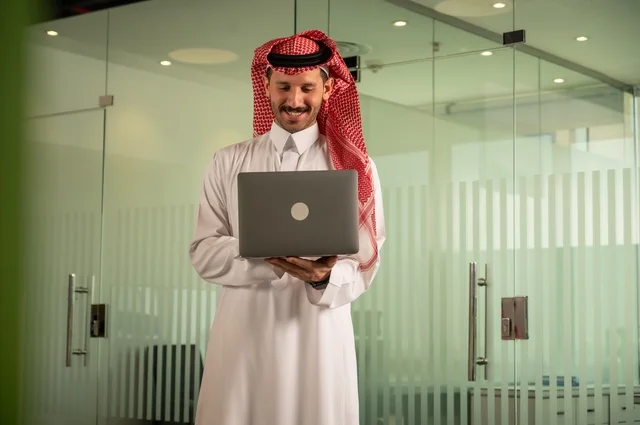 Management and organization of business affairs, using modern and advanced technology, an administrative office profession and job, an Arab Gulf Saudi man wearing traditional attire working on a laptop inside the office, companies and offices in the Kingdom of Saudi Arabia.
