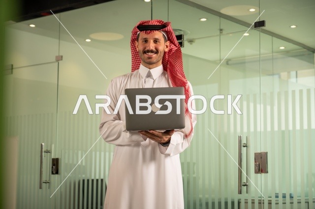 Companies and offices in the Kingdom of Saudi Arabia, managing and organizing business affairs, an administrative office profession and job, using a modern and advanced technical device, an Arab Gulf Saudi man wearing traditional attire and a shemagh working on a laptop inside the office.