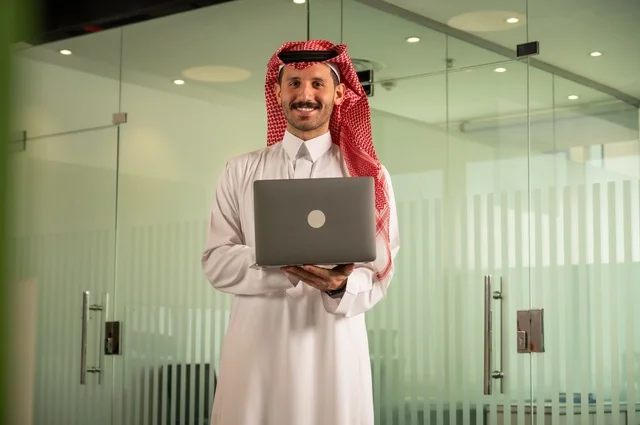 Companies and offices in the Kingdom of Saudi Arabia, managing and organizing business affairs, an administrative office profession and job, using a modern and advanced technical device, an Arab Gulf Saudi man wearing traditional attire and a shemagh working on a laptop inside the office.