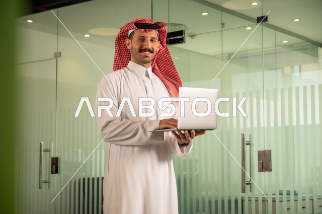 استخدام جهاز تقني حديث ومتطور، إدارة شؤون الأعمال وتنظيمها،  مهنة ووظيفة ادارية مكتبية، رجل عربي خليجي سعودي يرتدي الثوب التقليدي والشماغ يقوم بالعمل على الحاسوب المحمول داخل المكتب، الشركات والمكاتب في المملكة العربية السعودية