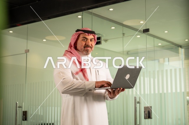 إدارة شؤون الأعمال وتنظيمها، استخدام جهاز تقني حديث ومتطور، مهنة ووظيفة ادارية مكتبية، رجل عربي خليجي سعودي يرتدي الثوب التقليدي يقوم بالعمل على الحاسوب المحمول داخل المكتب، الشركات والمكاتب في المملكة العربية السعودية