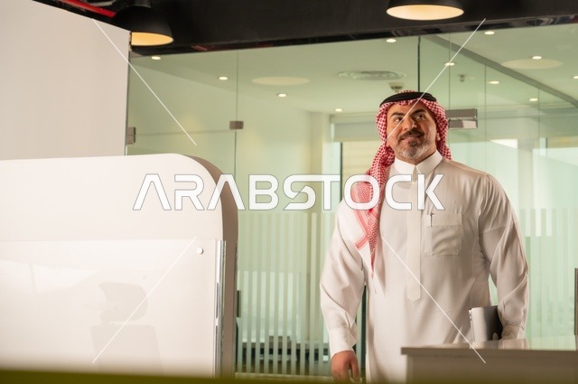 ايماءات الثقة والفخر، الشركات والمكاتب في المملكة العربية السعودية،  إدارة شؤون الأعمال وتنظيمها،  مهنة ووظيفة ادارية مكتبية، استخدام جهاز تقني حديث ومتطور، رجل عربي خليجي سعودي يرتدي الثوب التقليدي والشماغ يقف داخل المكتب
