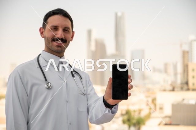 عرض شاشة سوداء فارغة عبر الهاتف الجوال، النظر إلى الكاميرا بإيماءات السرور، العمل في القطاعات الصحية، تسجيل التقارير والحالات الصحيه, طبيب عربي خليجي سعودي مبتسم يرتدي المعطف الأبيض يقف في الهواء الطلق، خلفية أبراج وناطحات سحاب في الرياض
