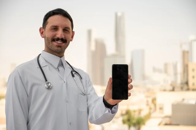 عرض شاشة سوداء فارغة عبر الهاتف الجوال، النظر إلى الكاميرا بإيماءات السرور، العمل في القطاعات الصحية، تسجيل التقارير والحالات الصحيه, طبيب عربي خليجي سعودي مبتسم يرتدي المعطف الأبيض يقف في الهواء الطلق، خلفية أبراج وناطحات سحاب في الرياض