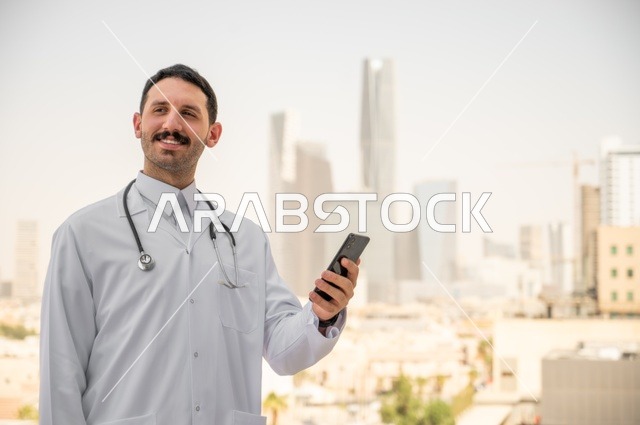النظر إلى الكاميرا بإيماءات السرور، العمل في القطاعات الصحية، تسجيل التقارير والحالات الصحيه, طبيب عربي خليجي سعودي مبتسم يرتدي المعطف الأبيض يقف في الهواء الطلق، خلفية أبراج وناطحات سحاب في الرياض، تصفح مواقع التواصل الاجتماعي