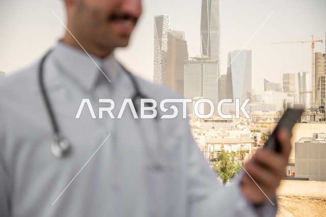 تسجيل الكشوفات والحالات، العمل في القطاعات الصحية، تسجيل التقارير والحالات الصحيه, صورة مقربة لطبيب عربي خليجي سعودي مبتسم يرتدي المعطف الأبيض يقف في الهواء الطلق، خلفية أبراج وناطحات سحاب في الرياض، تصفح مواقع التواصل الاجتماعي