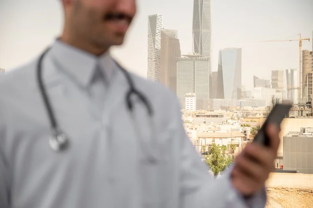 تسجيل الكشوفات والحالات، العمل في القطاعات الصحية، تسجيل التقارير والحالات الصحيه, صورة مقربة لطبيب عربي خليجي سعودي مبتسم يرتدي المعطف الأبيض يقف في الهواء الطلق، خلفية أبراج وناطحات سحاب في الرياض، تصفح مواقع التواصل الاجتماعي