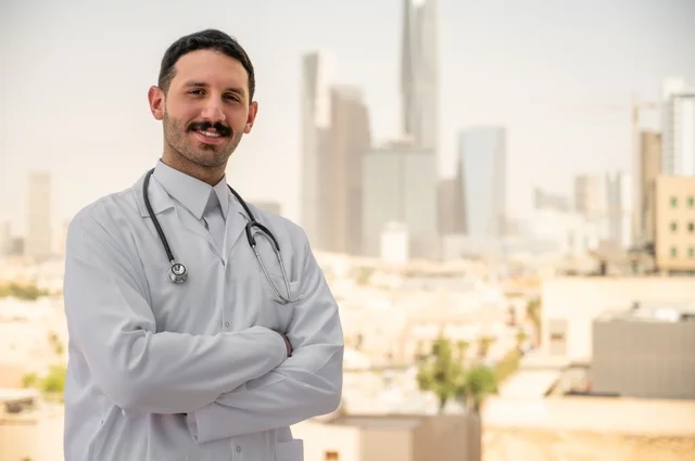 العمل في القطاعات الصحية، قياس معدل ضربات القلب من خلال سماعة الطبيب، تسجيل الكشوفات والحالات، تسجيل التقارير والحالات الصحيه, طبيب عربي خليجي سعودي مبتسم يرتدي المعطف الأبيض يقف في الهواء الطلق، خلفية أبراج وناطحات سحاب في الرياض