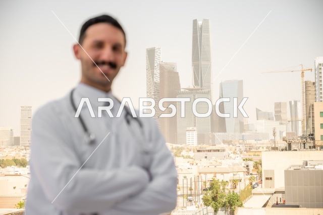 طبيب عربي خليجي سعودي مبتسم يرتدي المعطف الأبيض يقف في الهواء الطلق ايماءات ايدي متقاطعة، العمل في القطاعات الصحية، قياس معدل ضربات القلب من خلال سماعة الطبيب، تسجيل الكشوفات والحالات، تسجيل التقارير والحالات الصحيه, خلفية أبراج وناطحات سحاب في الرياض