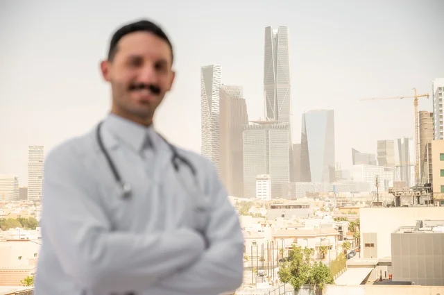 A smiling Saudi Arabian Gulf Arab doctor wearing a white coat stands outdoors with crossed arms, working in the health sectors, measuring heart rate with a stethoscope, recording examinations and cases, documenting reports and health conditions, with a background of towers and skyscrapers in Riyadh.