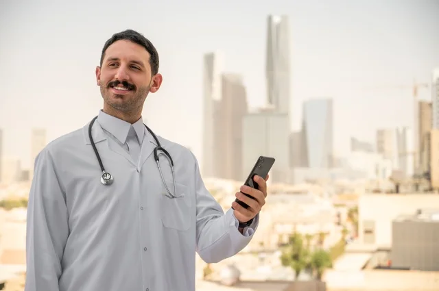 النظر إلى الكاميرا بإيماءات السرور، العمل في القطاعات الصحية، تسجيل التقارير والحالات الصحيه, طبيب عربي خليجي سعودي مبتسم يرتدي المعطف الأبيض يقف في الهواء الطلق، خلفية أبراج وناطحات سحاب في الرياض، تصفح مواقع التواصل الاجتماعي