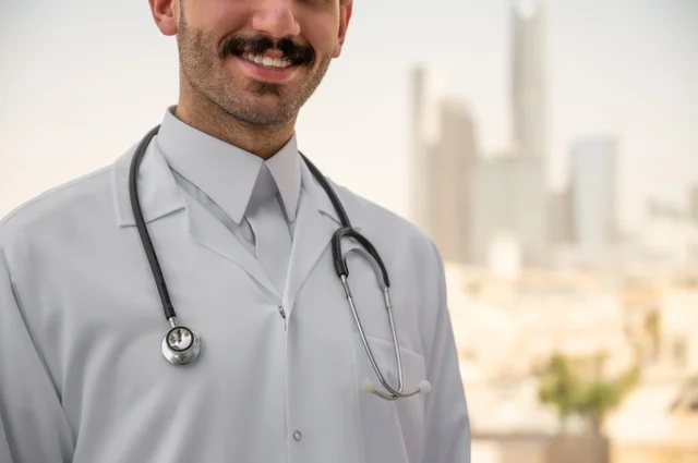قياس معدل ضربات القلب من خلال سماعة الطبيب، تسجيل الكشوفات والحالات، العمل في القطاعات الصحية، تسجيل التقارير والحالات الصحيه, صورة مقربة لطبيب عربي خليجي سعودي مبتسم يرتدي المعطف الأبيض يقف في الهواء الطلق، خلفية أبراج وناطحات سحاب في الرياض
