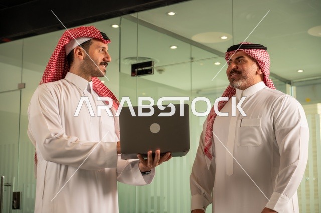 عقد صفقات عمل جديدة ناجحة بين رجال الأعمال،  مفهوم التعاون والعمل المشترك, صداقات وتعارف وبناء علاقات اجتماعية، صورة مقربة لرجلان سعوديان عربيان خليجيان يرتديان الزي التقليدي يقومون بالعمل على الحاسب المحمول، رجل اعمال يرتدي الشماغ و الثوب التقليدي
